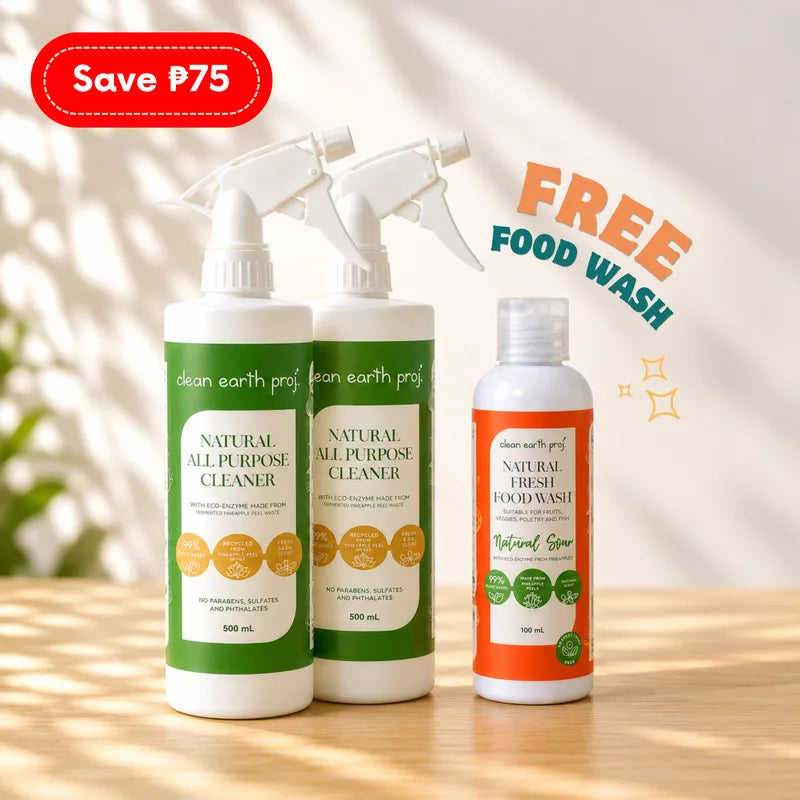 Three Clean Earth natural cleaning products set on a sunlit wooden table: two 500ml white spray bottles of Natural All-Purpose Cleaner in green packaging alongside a 100ml orange bottle of Natural Fresh Food Wash